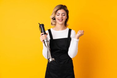 Sarı arka planda izole edilmiş el blenderi kullanan kız galibiyeti kutluyor.