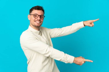 Mavi arka planda izole edilmiş beyaz tenli yakışıklı bir adam, parmağını yana uzattı ve bir ürün sundu.