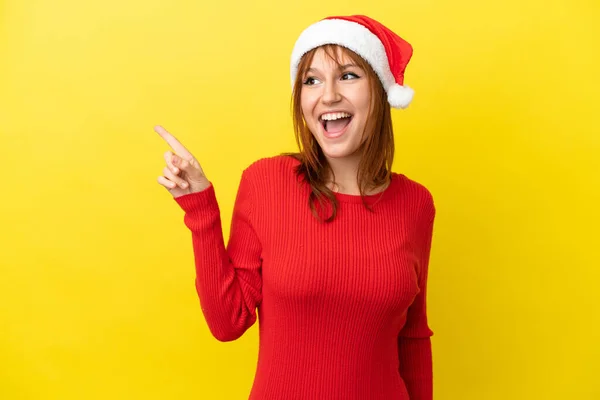 红头发姑娘戴着一顶黄色背景的圣诞帽 想在举起一只手指头的同时实现这个解决方法 — 图库照片