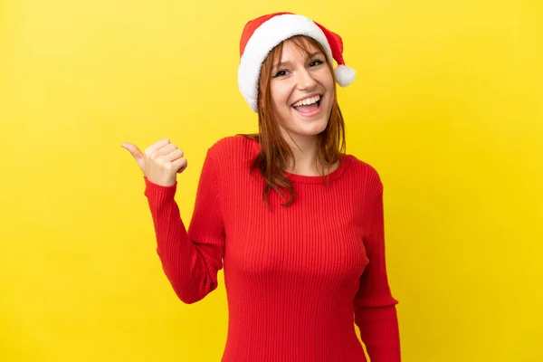 红头发姑娘 头戴一顶黄色背景的圣诞帽 指着侧面展示自己的产品 — 图库照片