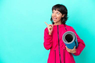 Mavi arka planda bir minder tutarak yoga derslerine giden genç kız çözümü bir parmağını kaldırarak bulmayı umuyor.