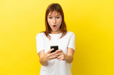 Kızıl saçlı kız sarı arka planda izole edilmiş. Kameraya bakarken şaşırmış bir ifadeyle cep telefonunu kullanıyor.