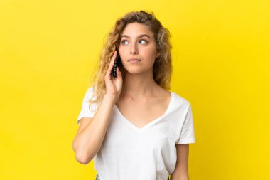 Sarı arka planda izole edilmiş cep telefonu kullanan genç sarışın kadın yan tarafa bakıyor.