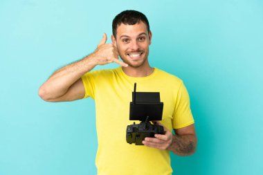 Brezilyalı bir adam uzaktan kumandayla izole edilmiş mavi arkaplanı kontrol ediyor ve telefon jesti yapıyor. Bana geri dönüş işareti ver.