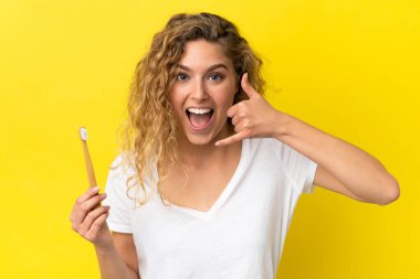Sarı arka planda izole edilmiş dişlerini fırçalayan beyaz bir kadın telefon jesti yapıyor. Bana geri dönüş işareti ver.