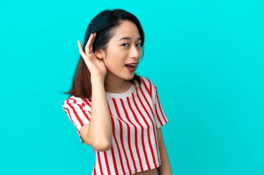 Genç Vietnamlı bir kadın mavi arka planda izole edilmiş bir şekilde elini kulağına koyarak bir şeyler dinliyor.