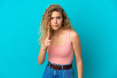 Mavi arka planda izole edilmiş genç sarışın kadın hayal kırıklığına uğramış ve ön tarafı işaret ediyor.