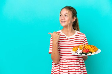 Mavi arka planda izole edilmiş waffle tutan küçük beyaz kız bir ürünü sunmak için yan tarafı işaret ediyor.