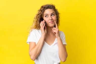 Sarı arka planda izole edilmiş cep telefonu kullanan genç sarışın kadın yukarı bakarken bir fikir düşünüyor.