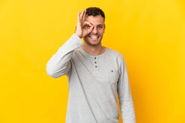 Sarı arka planda izole edilmiş beyaz, yakışıklı genç bir adam parmaklarıyla tamam işareti gösteriyor.