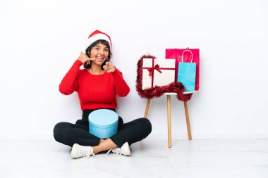 Genç bir kız Noel 'i kutluyor. Beyaz bir bakkal bahçesinde izole bir şekilde oturuyor. Telefon işareti yapıyor ve ön tarafı işaret ediyor.