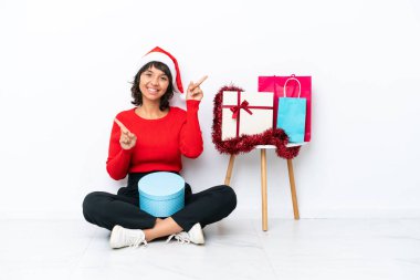 Noel 'i kutlayan genç bir kız, beyaz fırında izole bir şekilde yerde oturmuş, parmaklarını yan tarafları göstererek mutlu bir şekilde oturuyor.