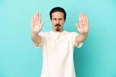 Mavi arka planda izole edilmiş genç beyaz adam dur hareketi yapıyor ve hayal kırıklığına uğruyor.