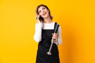 El blenderi kullanan kız sarı arkaplanda izole edilmiş cep telefonuyla sohbet ediyor.