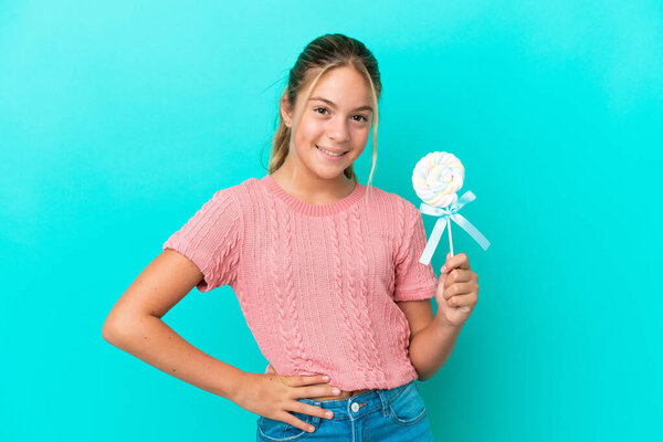 Little Caucasian girl holding a lollipop isolated on blue background posing with arms at hip and smiling