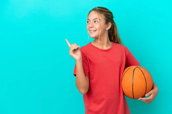 指を持ち上げながら解決策を実現しようとしている青の背景に隔離されたバスケットボールを再生する小さな白人の女の子 — ストック写真