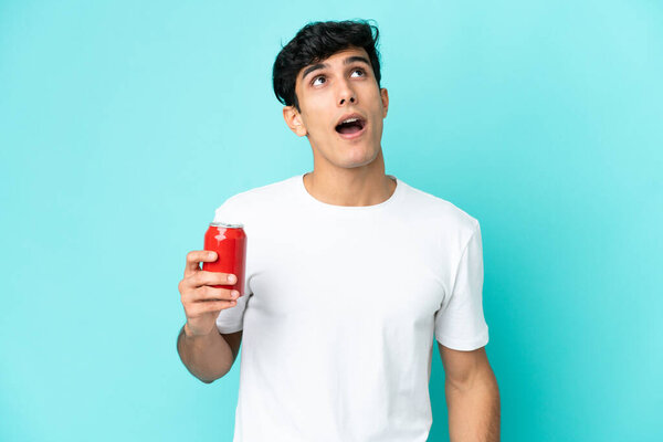 Young Argentinian man holding a refreshment isolated on blue background looking up and with surprised expression