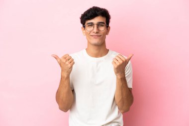 Arjantinli genç adam pembe arka planda izole edilmiş. Baş parmakları havada ve gülümsüyor.
