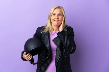 Elinde motosiklet kaskı olan orta yaşlı bir kadın. Mor arka planda izole edilmiş, şaşırmış ve şok olmuş bir yüz ifadesi var.