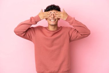 Arjantinli genç bir adam pembe arka planda izole edilmiş. Gözlerini elleriyle kapatıyor ve gülümsüyor.