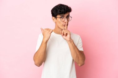 Pembe arka planda izole edilmiş genç Arjantinli bir adam kenarı işaret ediyor ve sessizlik hareketi yapıyor.