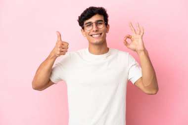 Pembe arka planda izole edilmiş genç Arjantinli adam işaret ve baş parmak hareketi gösteriyor.