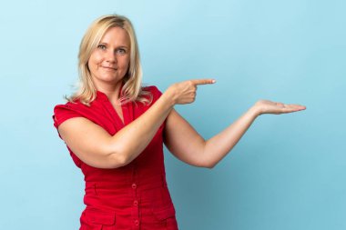 Mavi arka planda izole edilmiş orta yaşlı bir kadın, bir ilan vermek için avucunun içinde hayali bir alan tutuyor.