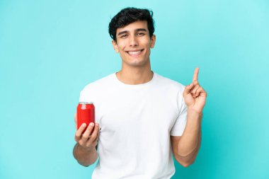 Arjantinli genç adam mavi arka planda izole edilmiş bir içecek tutuyor ve en iyinin işareti olarak parmağını kaldırıyor.