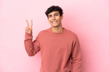 Pembe arka planda izole edilmiş genç Arjantinli adam gülümsüyor ve zafer işareti gösteriyor.