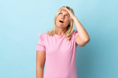 Mavi arka planda soyutlanmış orta yaşlı bir kadın ön tarafa bakarken sürpriz jest yapıyor.