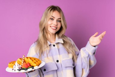 Mor arka planda izole edilmiş waffle 'ları tutan genç Rus kız davet etmek için ellerini yana uzatıyor.