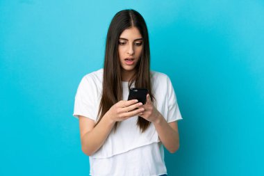 Mavi arka planda izole edilmiş genç beyaz kadın kameraya bakarken şaşırmış bir ifadeyle cep telefonunu kullanıyor.