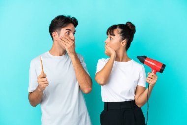 Mavi arka planda saç kurutma makinesi ve diş fırçası tutan genç ve melez bir çift uygunsuz bir şey söyledikleri için ağızlarını elleriyle kapatıyor.