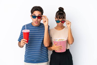Beyaz arka planda soyutlanmış genç melez arkadaşlar 3D gözlüklü ve elinde koca bir kova patlamış mısırla şaşırdılar.