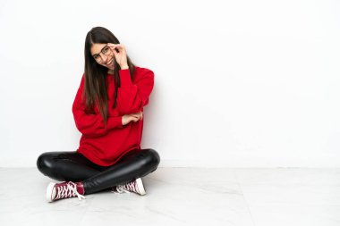 Genç bir kadın beyaz arka planda gözlüklü ve mutlu bir şekilde yerde oturuyor.