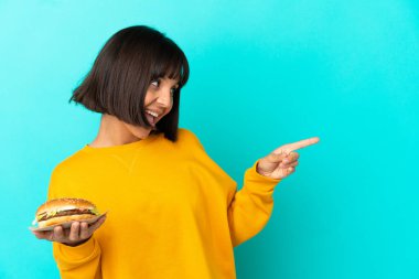 Genç esmer kadın izole edilmiş arka planda bir burger tutuyor. Parmaklarını kenarı gösteriyor ve bir ürün sunuyor.