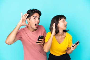Mavi arka planda cep telefonu tutan melez bir çift kulağına bir şey dayayıp onu dinliyor.