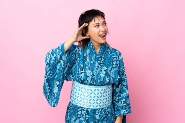 İzole edilmiş mavi arka planda kimono giyen genç bir kadın kulağına elini sürerek bir şeyler dinliyor.