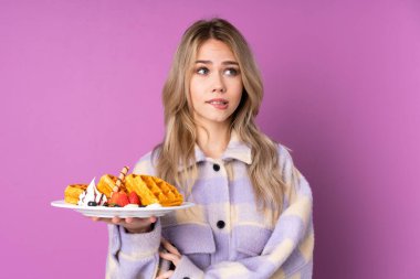 Elinde waffle tutan genç bir Rus kız. Mor arka planda izole edilmiş ve kafası karışık bir yüz ifadesi var.