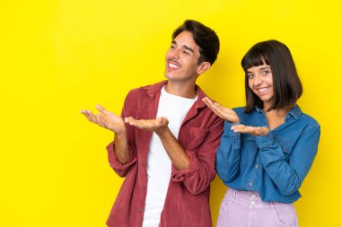 Sarı arka planda soyutlanmış genç melez bir çift davet etmek için ellerini yana uzatıyor.