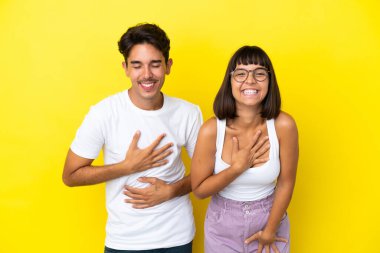 Sarı arka planda izole edilmiş genç melez çift ellerini göğsüne koyup gülümsüyor.