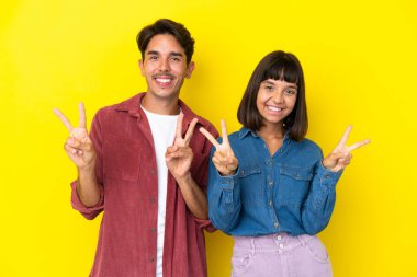 Sarı arka planda izole edilmiş genç melez çift gülümsüyor ve zafer işareti gösteriyorlar.