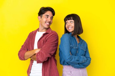 Sarı arka planda izole edilmiş genç ve melez bir çift gülümseyerek omuzlarının üzerinden bakıyor.