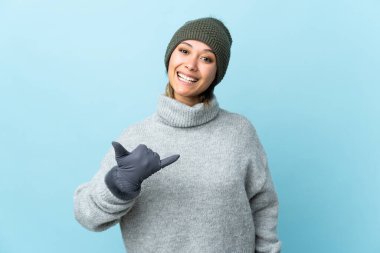Kışlık şapkalı genç Uruguaylı kız mavi arka planda izole edilmiş bir şekilde telefon jesti yapıyor.