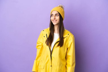 Mor arka planda izole edilmiş, yağmura dayanıklı bir ceket giyen genç beyaz kadın yukarı bakarken bir fikir düşünüyor.