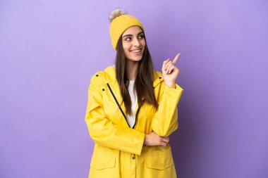 Mor arka planda izole edilmiş, yağmura dayanıklı bir ceket giyen beyaz bir kadın harika bir fikre işaret ediyor.