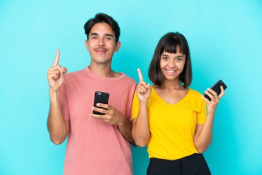 Mavi arka planda cep telefonu tutan genç melez çift en iyinin işareti olarak parmağını kaldırıyor.