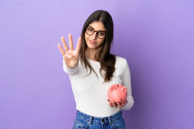 Mor arka planda izole edilmiş domuz kumbarası tutan beyaz bir kadın mutlu ve parmakları olan dört kişi sayıyor.