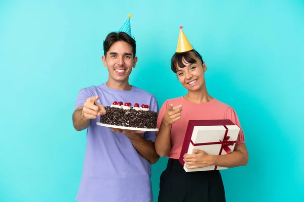 年轻的混血种族夫妇拿着生日蛋糕 带着自信的表情用蓝色的背景手指指着你 — 图库照片