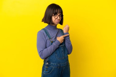 Genç hamile kadın izole edilmiş sarı arka planda geç kalma jesti yapıyor.